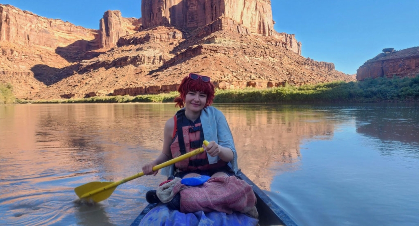 A person sits at the back of a canoe using a yellow paddle. Beyond the water behind them there is a large red rock formation. 